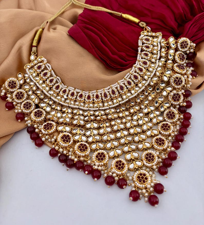 May include: A gold-toned statement necklace with intricate detailing. The necklace features a layered design with red and white accents, including small pearls and red beads. The necklace is displayed on a neutral background.