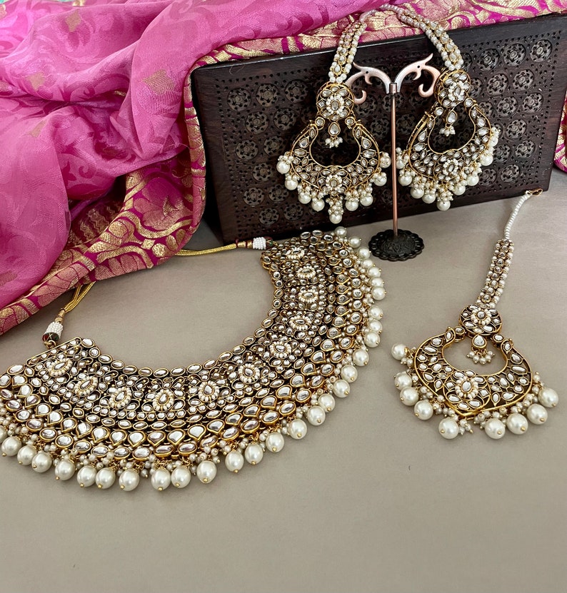 Pu&ograve; includere: Un set di gioielli da sposa in oro e bianco con una collana, orecchini e un tikka. La collana &egrave; di tipo choker con pi&ugrave; strati di perle e pietre. Gli orecchini sono lunghi e pendenti con perle e pietre. Il tikka &egrave; un piccolo copricapo con perle e pietre.