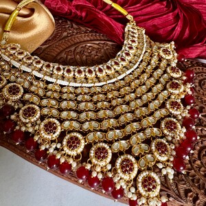 May include: A gold-toned necklace with intricate detailing, featuring red and white accents. The necklace has multiple layers of ornate pendants and dangling red beads. The necklace is displayed on a wooden surface with a red fabric backdrop.