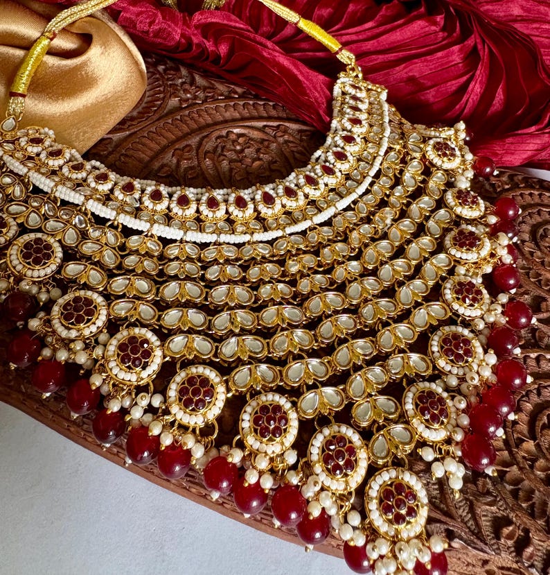 May include: A gold-toned necklace with intricate detailing, featuring red and white accents. The necklace has multiple layers of ornate pendants and dangling red beads. The necklace is displayed on a wooden surface with a red fabric backdrop.