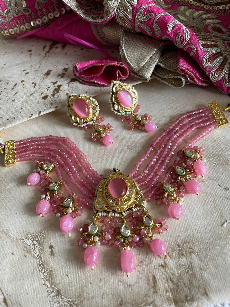 May include: A pink and gold layered beaded necklace with a large pink stone pendant and matching earrings. The necklace has multiple strands of pink beads and a gold pendant with a pink stone in the center. The earrings are also pink and gold with a pink stone in the center.