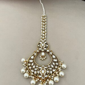 Pu&ograve; includere: Un maang tikka, un copricapo tradizionale indiano, in oro e perle bianche, con un grande design circolare decorato e una catena di perle.
