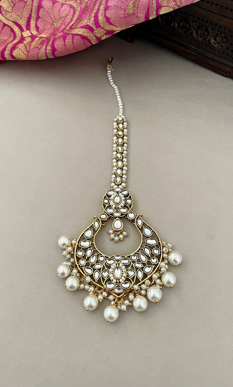 Pu&ograve; includere: Un maang tikka, un copricapo tradizionale indiano, in oro e perle bianche, con un grande design circolare decorato e una catena di perle.