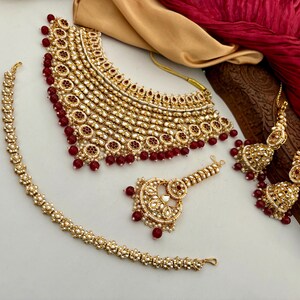 May include: A gold-toned jewelry set featuring a necklace, earrings, bracelet, and headpiece. The set is adorned with small, round, red beads and clear, reflective stones. The necklace and earrings have a layered design.