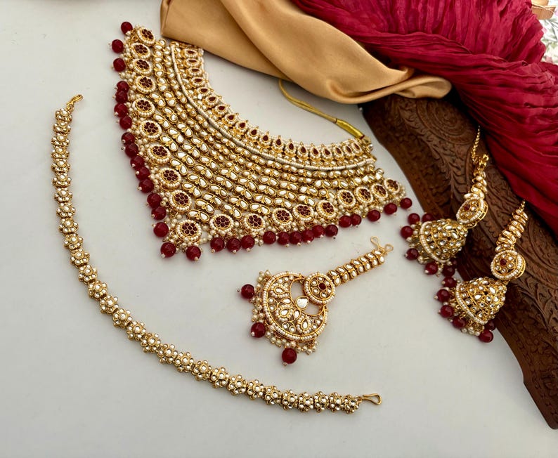 May include: A gold-toned jewelry set featuring a necklace, earrings, bracelet, and headpiece. The set is adorned with small, round, red beads and clear, reflective stones. The necklace and earrings have a layered design.