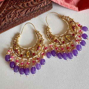 May include: A pair of gold-coloured hoop earrings with detailed patterns. The earrings have a crescent shape with red and white accents, and are decorated with hanging purple beads and pink and white beads.