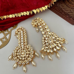 May include: A pair of gold earrings with pearl accents. The earrings are decorated with intricate designs and white stones.