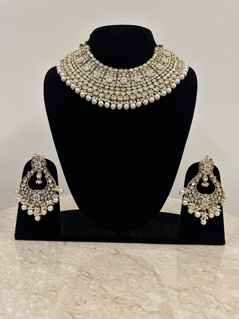 Pu&ograve; includere: Un set da sposa di collana e orecchini in oro e bianco con accenti di perle. La collana &egrave; un design multistrato con un grande ciondolo rotondo al centro. Gli orecchini sono a forma di lacrima con un design simile a quello della collana.