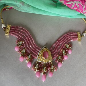 May include: A pink beaded choker necklace with a gold pendant featuring a pink stone. The necklace has multiple strands of beads and a dangling design with pink and white stones.