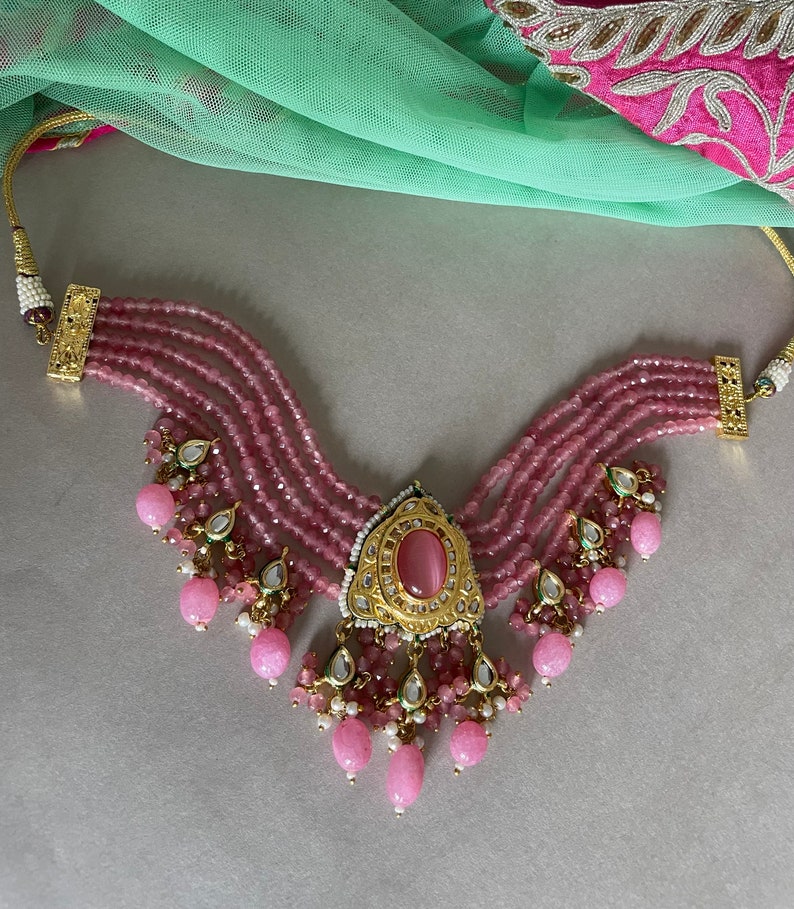 May include: A pink beaded choker necklace with a gold pendant featuring a pink stone. The necklace has multiple strands of beads and a dangling design with pink and white stones.