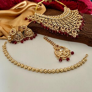 May include: A gold-toned bridal jewelry set featuring a necklace, earrings, a headpiece, and a bracelet. The set is adorned with white stones and red beads. The necklace has a bib design, and the earrings are bell-shaped.