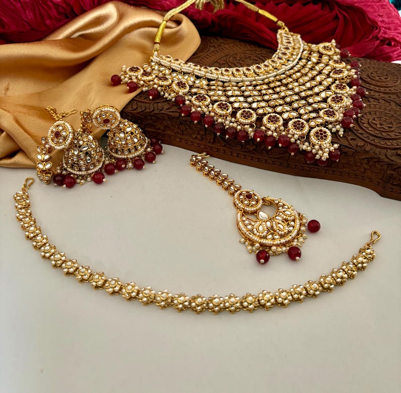 May include: A gold-toned bridal jewelry set featuring a necklace, earrings, a headpiece, and a bracelet. The set is adorned with white stones and red beads. The necklace has a bib design, and the earrings are bell-shaped.