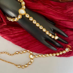 May include: A gold-tone hand and finger jewelry set with clear teardrop-shaped stones. The set includes a bracelet and a ring.