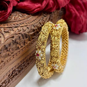 Peut inclure: Deux bracelets dorés ornés de motifs floraux et de feuilles, rehaussés de petites pierres précieuses rouges et blanches. Les bracelets sont présentés sur un fond blanc, avec une boîte en bois sculpté et un tissu rouge drapé en arrière-plan.