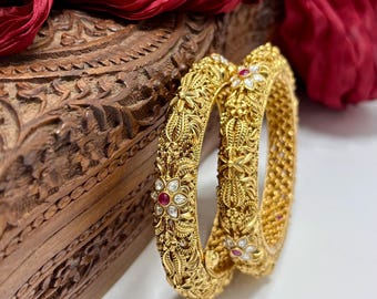 Puede incluir: Dos brazaletes dorados ornamentados con patrones florales y de hojas, acentuados con pequeñas piedras preciosas rojas y blancas. Los brazaletes se exhiben sobre una superficie blanca con una caja de madera tallada y una tela roja drapeada en el fondo.