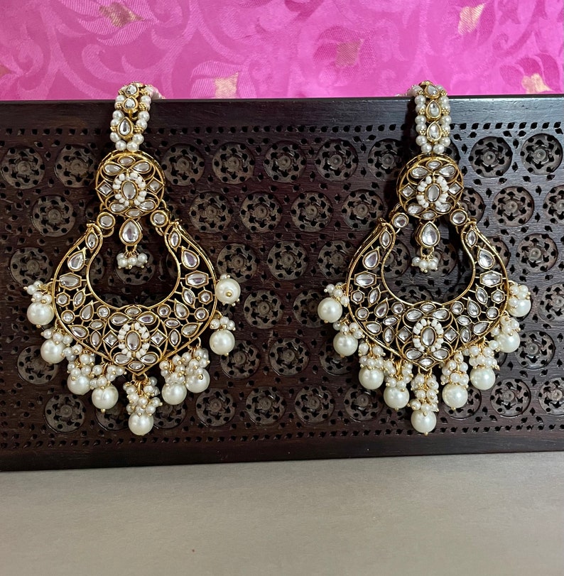 Pu&ograve; includere: Un paio di orecchini dorati con accenti di perle bianche. Gli orecchini presentano un design grande e ornato con dettagli intricati e una goccia di perla pendente.