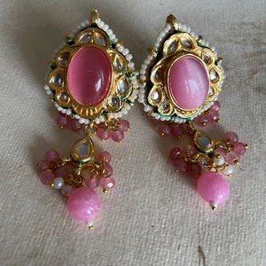 May include: A pair of pink and gold earrings with a teardrop design. The earrings feature a large pink stone surrounded by smaller stones and beads.