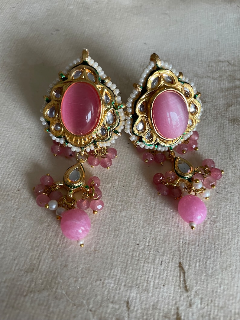 May include: A pair of pink and gold earrings with a teardrop design. The earrings feature a large pink stone surrounded by smaller stones and beads.