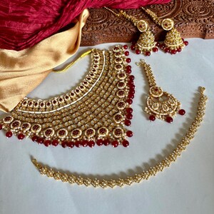 May include: A gold-toned jewelry set featuring a necklace, earrings, a headpiece, and a bracelet. The set is adorned with red and white accents. A red fabric and a wooden carved box are in the background.