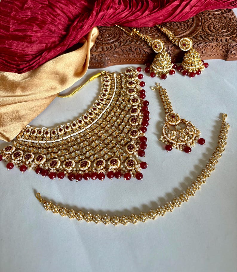 May include: A gold-toned jewelry set featuring a necklace, earrings, a headpiece, and a bracelet. The set is adorned with red and white accents. A red fabric and a wooden carved box are in the background.