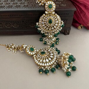 May include: A pair of gold-tone earrings with intricate designs and green and white stones. The earrings have a round shape and are adorned with pearls and beads.