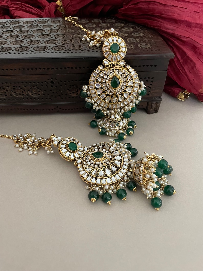 May include: A pair of gold-tone earrings with intricate designs and green and white stones. The earrings have a round shape and are adorned with pearls and beads.