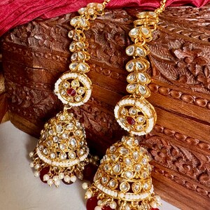 May include: Gold-toned chandelier earrings with intricate detailing. The earrings feature a bell-shaped design with clear and red accents, and small white pearls. The earrings are displayed on a wooden surface with a red fabric backdrop.