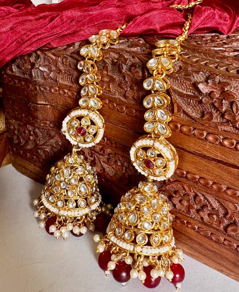 May include: Gold-toned chandelier earrings with intricate detailing. The earrings feature a bell-shaped design with clear and red accents, and small white pearls. The earrings are displayed on a wooden surface with a red fabric backdrop.