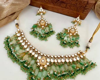 May include: A gold-toned necklace and earring set featuring green and white gemstones. The necklace has a bib design with a teardrop pendant and a fringe of green beads. The earrings mirror the necklace design, with a floral top and dangling green beads.