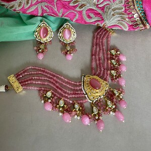 May include: A pink and gold layered necklace with a large pink stone pendant and matching earrings. The necklace features multiple strands of pink beads and intricate gold detailing.