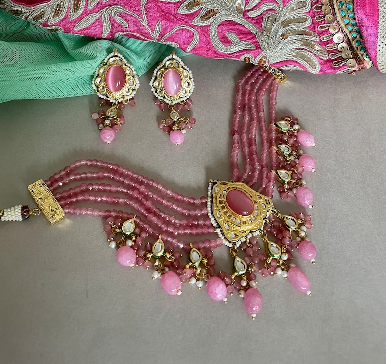 May include: A pink and gold layered necklace with a large pink stone pendant and matching earrings. The necklace features multiple strands of pink beads and intricate gold detailing.