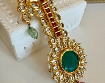 Könnte beinhalten: Aufwendiges goldfarbenes Schmuckstück mit einem großen ovalen grünen Edelstein in der Mitte, umgeben von kleineren klaren Steinen. Mit roten und klaren Akzenten sowie hängenden grünen und rosa Edelsteinen. Ein detailliertes, auffälliges Accessoire.
