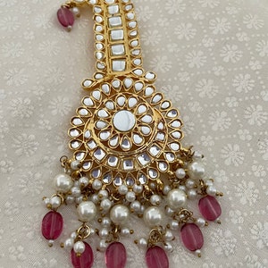 Op de afbeelding: Een sierlijk gouden haaraccessoire met een gebogen bovenkant en een cirkelvormige hanger. Het is versierd met heldere, vierkante en druppelvormige stenen, witte parels en roze kralen. De achtergrond is een crèmekleurige stof.