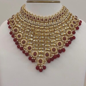May include: A gold-toned necklace with intricate detailing, featuring red and white accents. The necklace has a layered design with circular and teardrop-shaped elements, and is displayed on a white bust. The necklace is a statement piece.