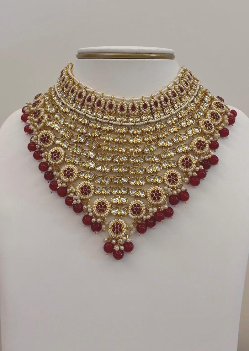 May include: A gold-toned necklace with intricate detailing, featuring red and white accents. The necklace has a layered design with circular and teardrop-shaped elements, and is displayed on a white bust. The necklace is a statement piece.