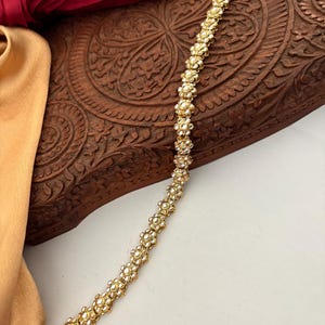 May include: A gold-toned, floral-patterned hair accessory. The piece is adorned with small, round, white gemstones, creating a delicate and ornate design. The accessory is likely for special occasions.