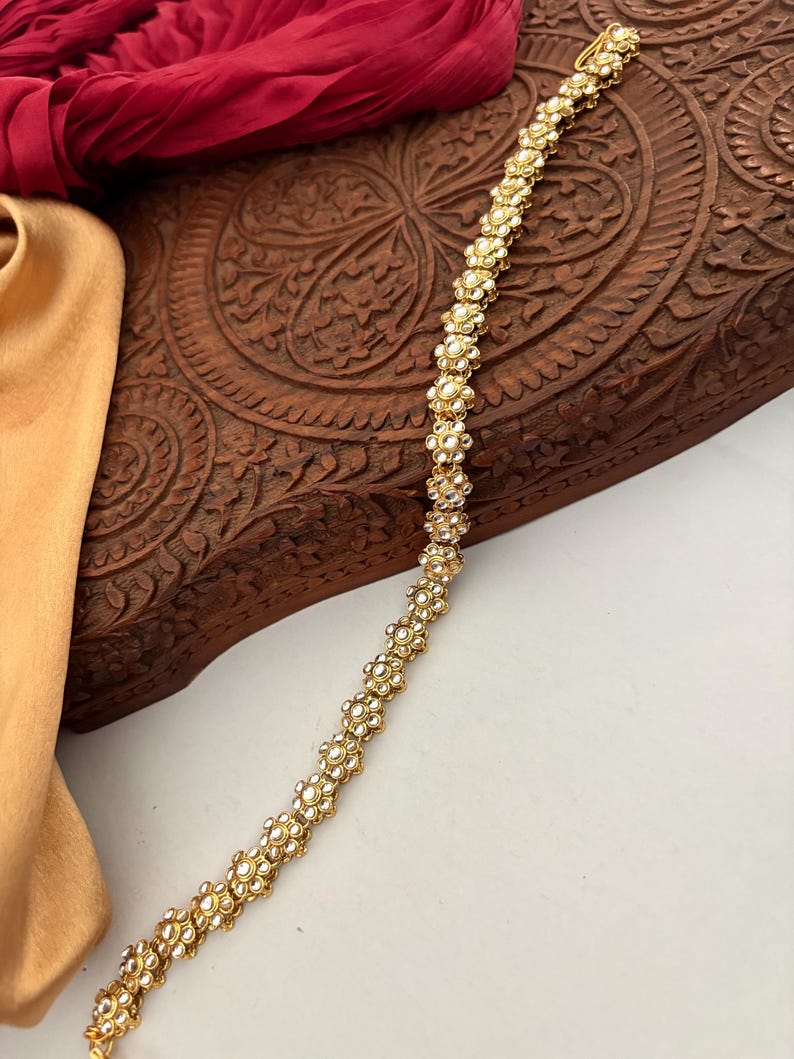 May include: A gold-toned, floral-patterned hair accessory. The piece is adorned with small, round, white gemstones, creating a delicate and ornate design. The accessory is likely for special occasions.