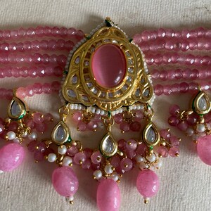 May include: A pink and gold beaded necklace with a large pink stone pendant. The pendant is surrounded by smaller stones and has a detailed design.