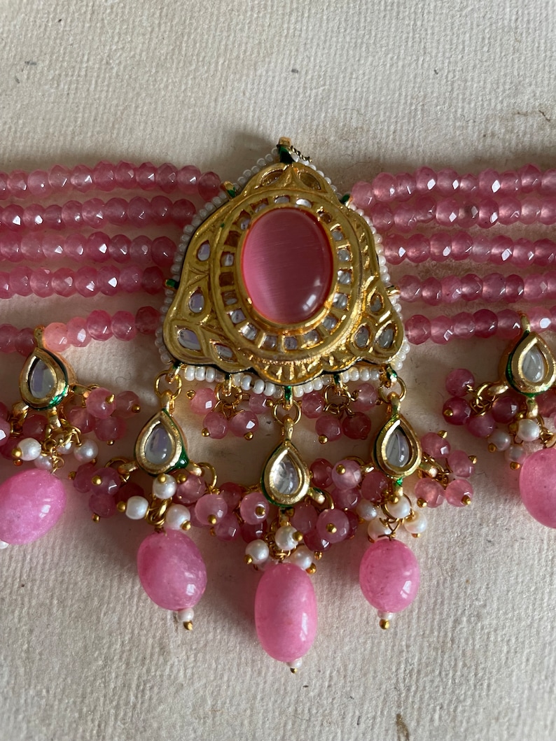 May include: A pink and gold beaded necklace with a large pink stone pendant. The pendant is surrounded by smaller stones and has a detailed design.