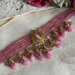 May include: A pink beaded choker necklace with a gold pendant featuring a pink stone. The necklace has multiple strands of beads and dangling pink and white beads.