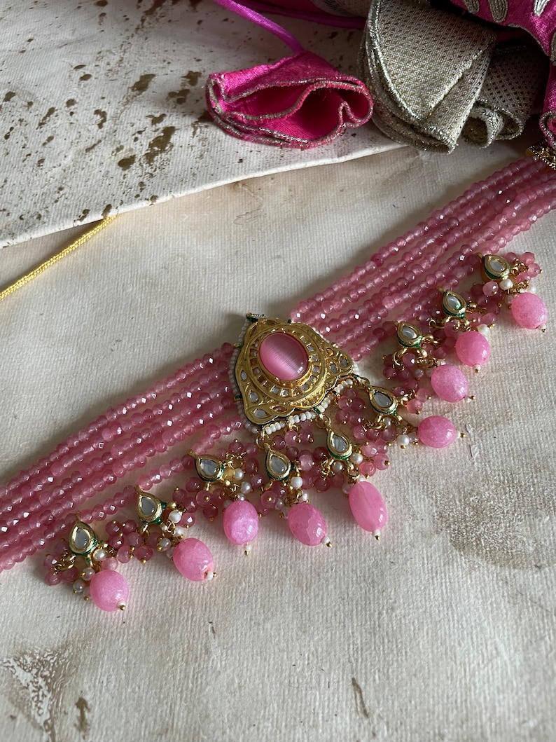 May include: A pink beaded choker necklace with a gold pendant featuring a pink stone. The necklace has multiple strands of beads and dangling pink and white beads.