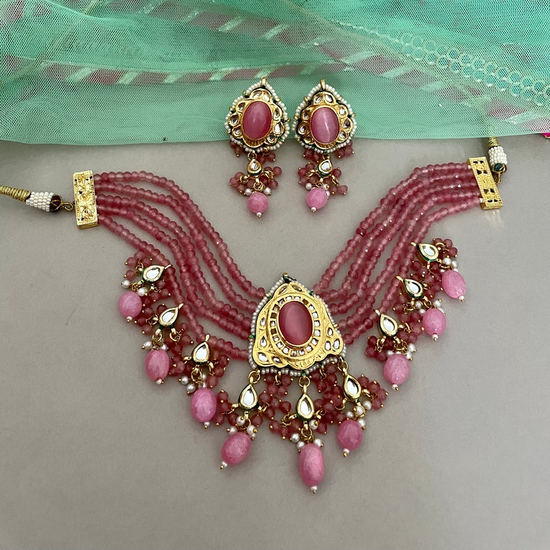 May include: A pink and gold layered necklace with a large pink stone pendant and matching earrings. The necklace has multiple strands of pink beads and the pendant is decorated with intricate gold details.