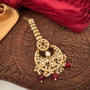 May include: A gold-toned headpiece with a crescent moon design, adorned with white and red gemstones and small pearls. The jewelry piece is set against a dark wooden surface and a red fabric.