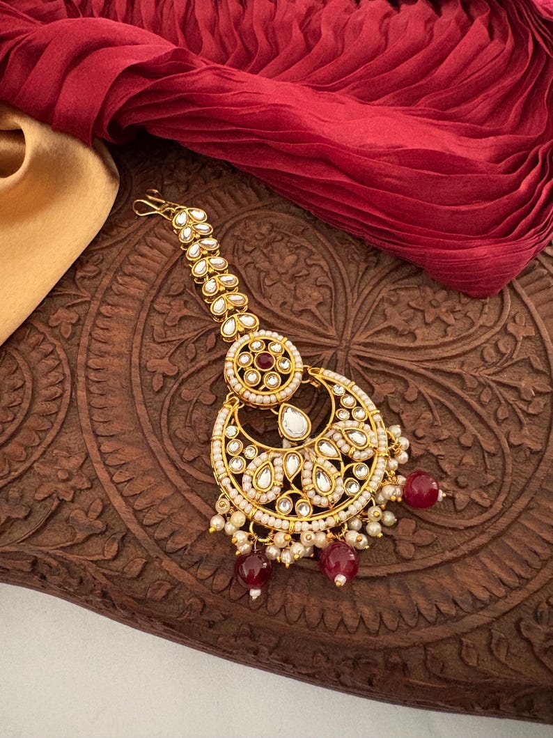 May include: A gold-toned headpiece with a crescent moon design, adorned with white and red gemstones and small pearls. The jewelry piece is set against a dark wooden surface and a red fabric.