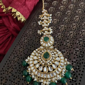 May include: A gold and white maang tikka with green stones and pearl accents. The maang tikka is a traditional Indian head ornament.