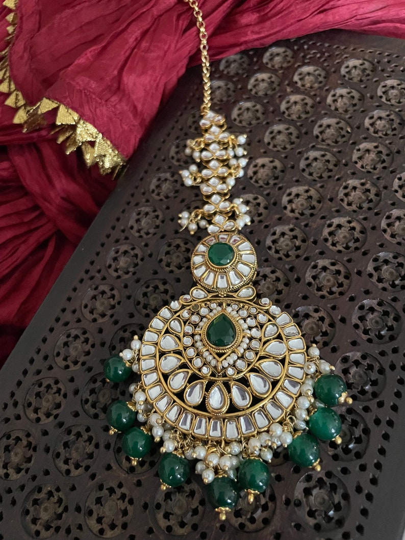 May include: A gold and white maang tikka with green stones and pearl accents. The maang tikka is a traditional Indian head ornament.