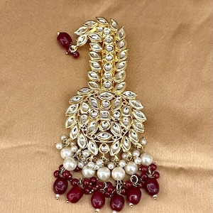 Peut inclure: Une ornement de cheveux doré avec un design en forme de larme, orné de pierres claires et de perles. L'ornement présente une chaîne pendante de perles rouges et blanches.