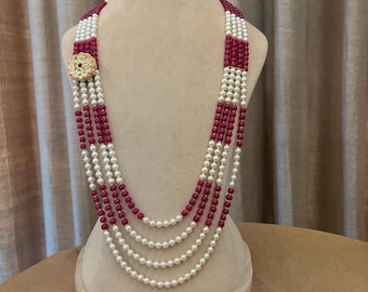May include: A multi-strand necklace with alternating sections of white pearls and deep red beads. The necklace features a decorative gold-toned clasp with a floral design. Additional gold beads are scattered on the surface below the necklace.