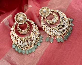 May include: A pair of gold-toned, ornate earrings. Each earring features a circular top, a crescent-shaped middle, and a larger, openwork bottom. The earrings are embellished with white stones, small pearls, and dangling green beads. The earrings rest on a pink fabric.