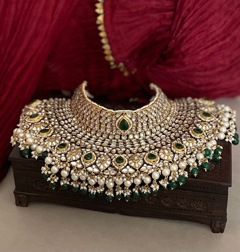 May include: A large, ornate gold necklace with white and green stones. The necklace is on a dark wooden box.